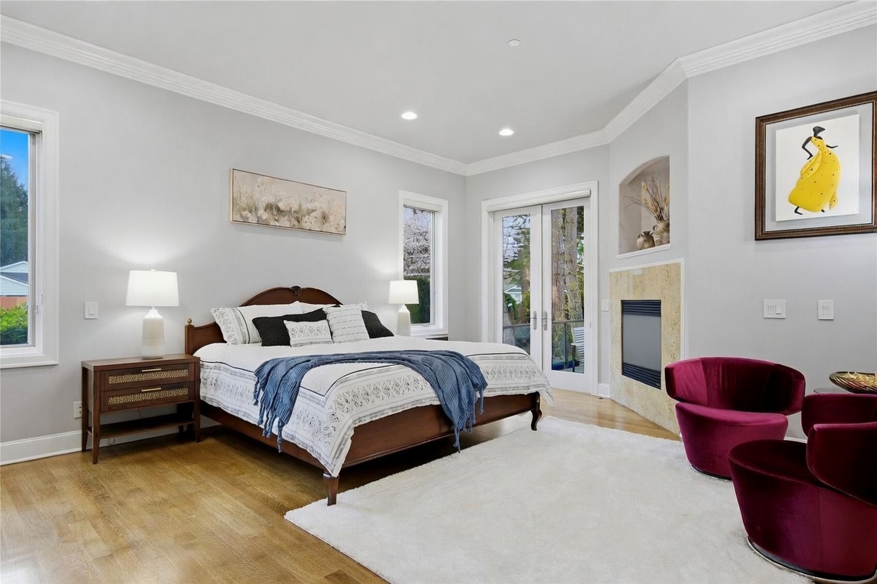 This spacious primary bedroom features light-toned walls, hardwood flooring, and a large white area rug that anchors the space. A dark wood bed frame is centered against the wall, flanked by matching nightstands and lamps, while a cozy seating area with two deep-red velvet chairs sits near a stone-clad fireplace. Large glass doors provide natural light and access to an outdoor space, creating an inviting and serene atmosphere.