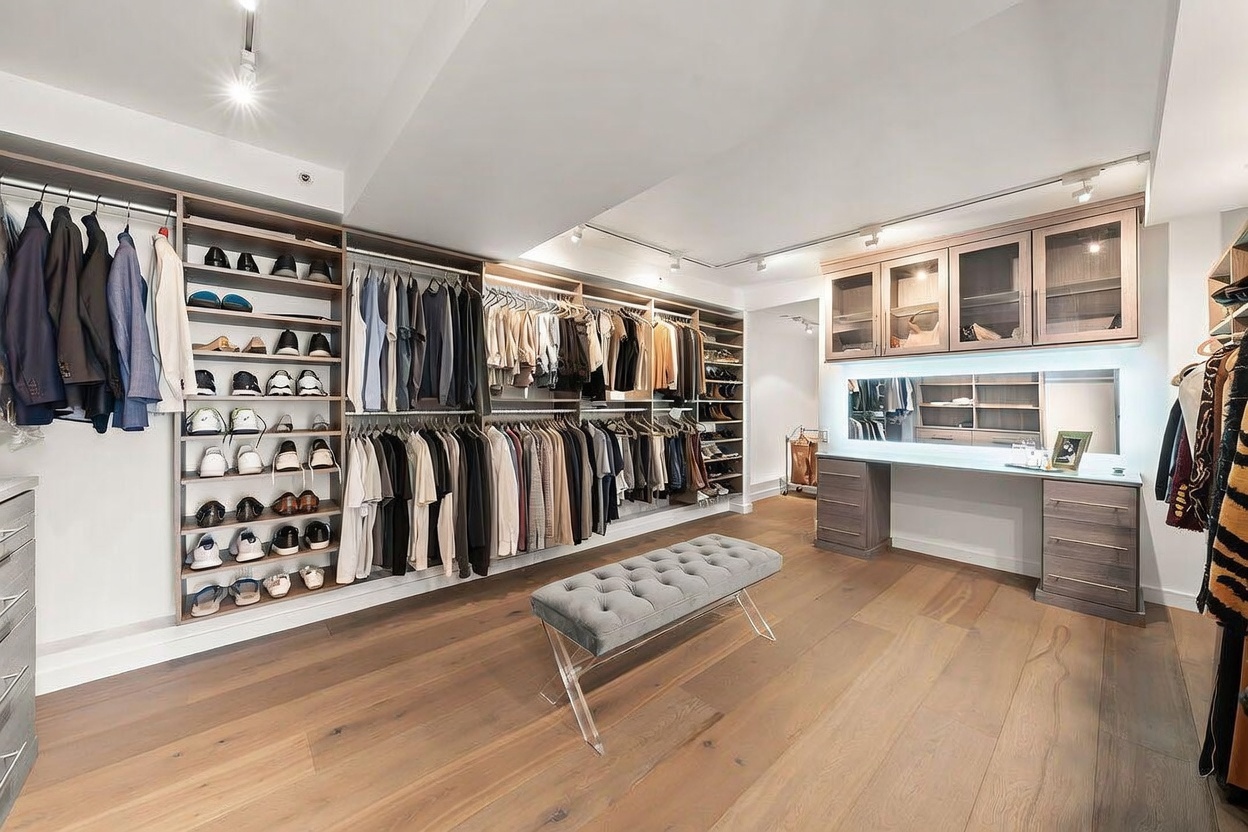 This expansive, well-organized walk-in closet features extensive custom cabinetry with open shelving, hanging rods, and a dedicated vanity area with a mirror and drawers. A tufted grey bench sits in the center of the room on light wood flooring, providing a functional and elegant focal point. The space is illuminated by recessed lighting and track lighting, creating a bright and sophisticated atmosphere.