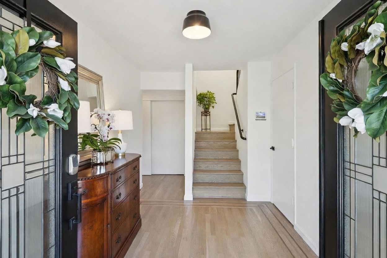 This bright and welcoming entryway features light wood flooring and a classic staircase leading to the upper level. A dark wood dresser with a mirror and floral arrangement sits to the left, while a black-framed glass door with a decorative wreath creates a striking focal point. The space is illuminated by a modern dome-style ceiling light, providing a clean and inviting first impression of the home.