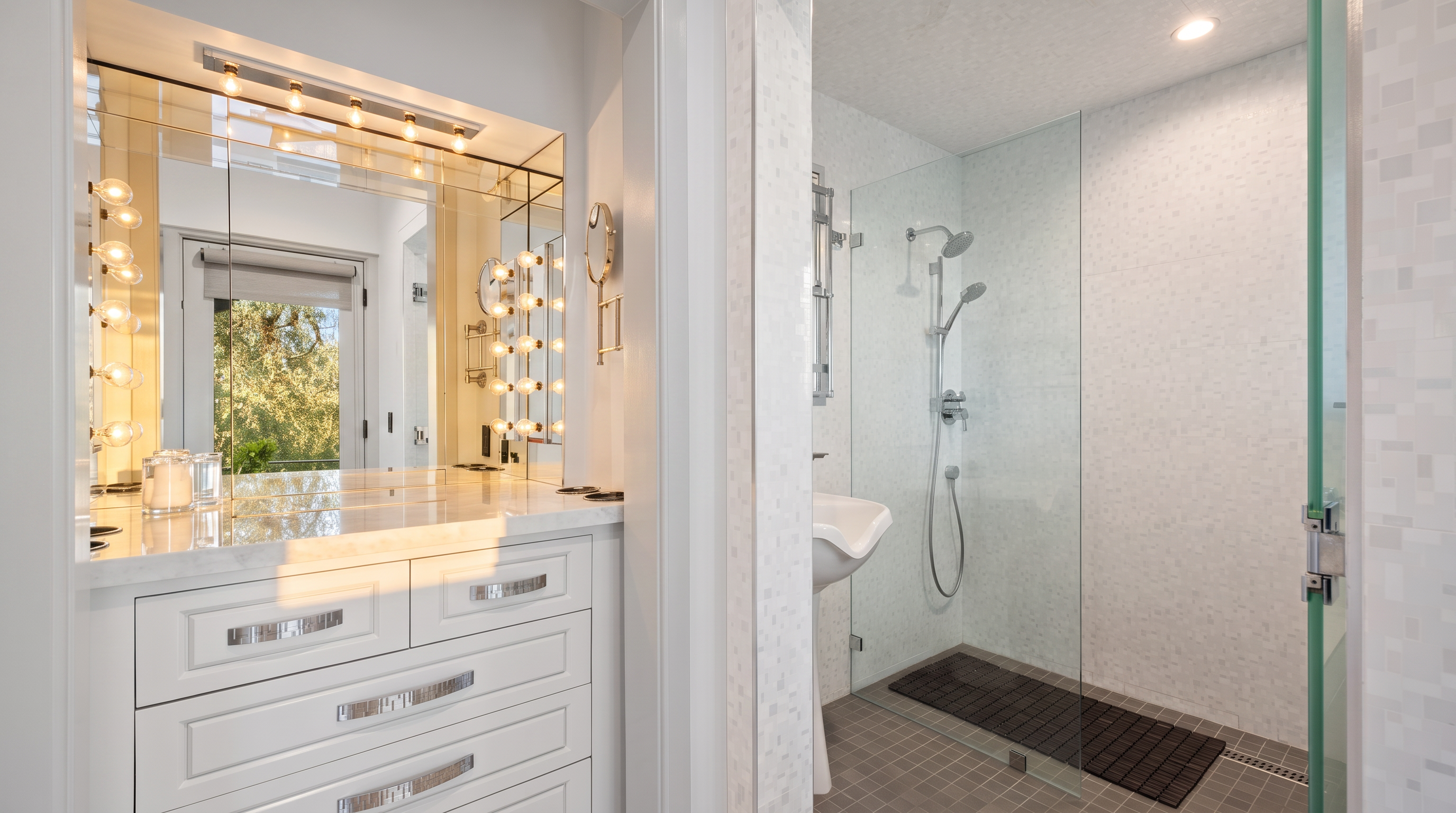 This primary bathroom features a clean, white vanity with polished hardware and a mirrored wall that reflects the adjacent space. To the right, a glass-enclosed walk-in shower is finished with light-colored wall tiles and a dark, textured floor mat. The perspective captures the transition between the vanity area and the shower, highlighting a bright, modern aesthetic.