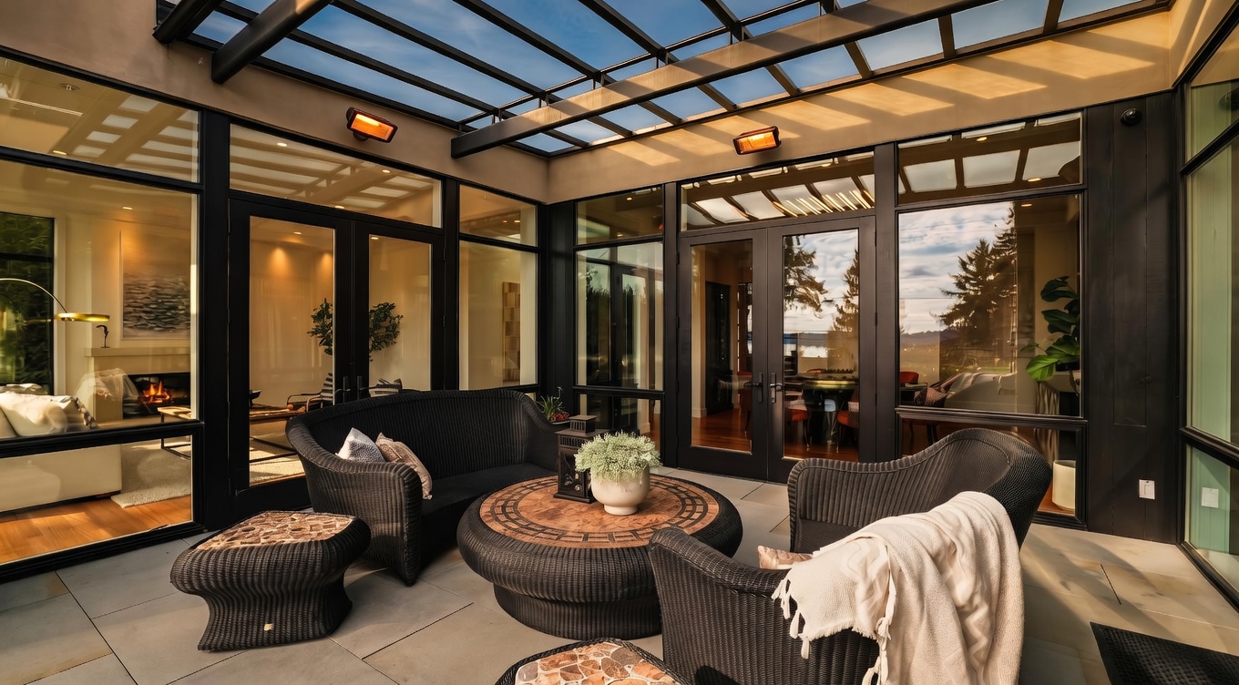 This covered outdoor patio features a sleek, modern glass-paneled roof supported by a dark metal frame, creating a bright and airy atmosphere. The space is furnished with dark wicker seating, including a sofa, armchairs, and a round stone-topped coffee table, all set upon large, neutral-toned stone pavers. Large glass walls and doors provide a seamless transition to the interior living spaces, while two wall-mounted heaters ensure comfort in this sophisticated, sheltered lounge area.
