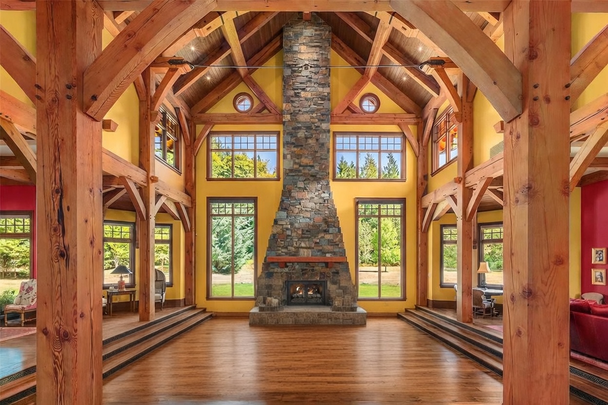 This grand living room features a dramatic vaulted ceiling with exposed timber trusses and a prominent stone-clad chimney breast that serves as the room's focal point. The space is anchored by a large blue sofa and a central console table, all set upon a patterned area rug atop rich wood flooring. Tall windows on either side of the fireplace provide abundant natural light and views of the surrounding landscape, creating an airy and sophisticated atmosphere.
