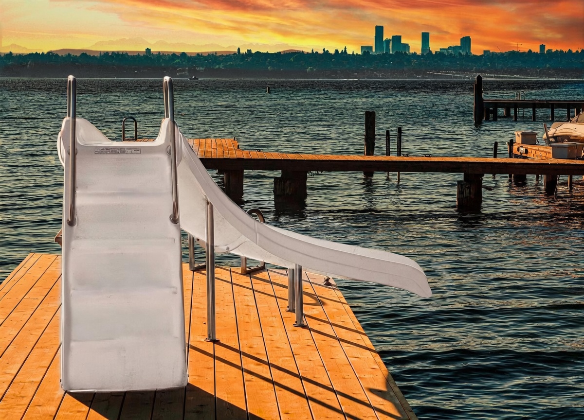 This image captures a serene waterfront scene from a wooden dock, featuring a white slide that leads directly into the water. In the background, a stunning sunset illuminates the horizon, casting a warm glow over the distant city skyline and mountain silhouettes. The perspective is low and centered, emphasizing the peaceful transition from the private dock to the expansive lake.