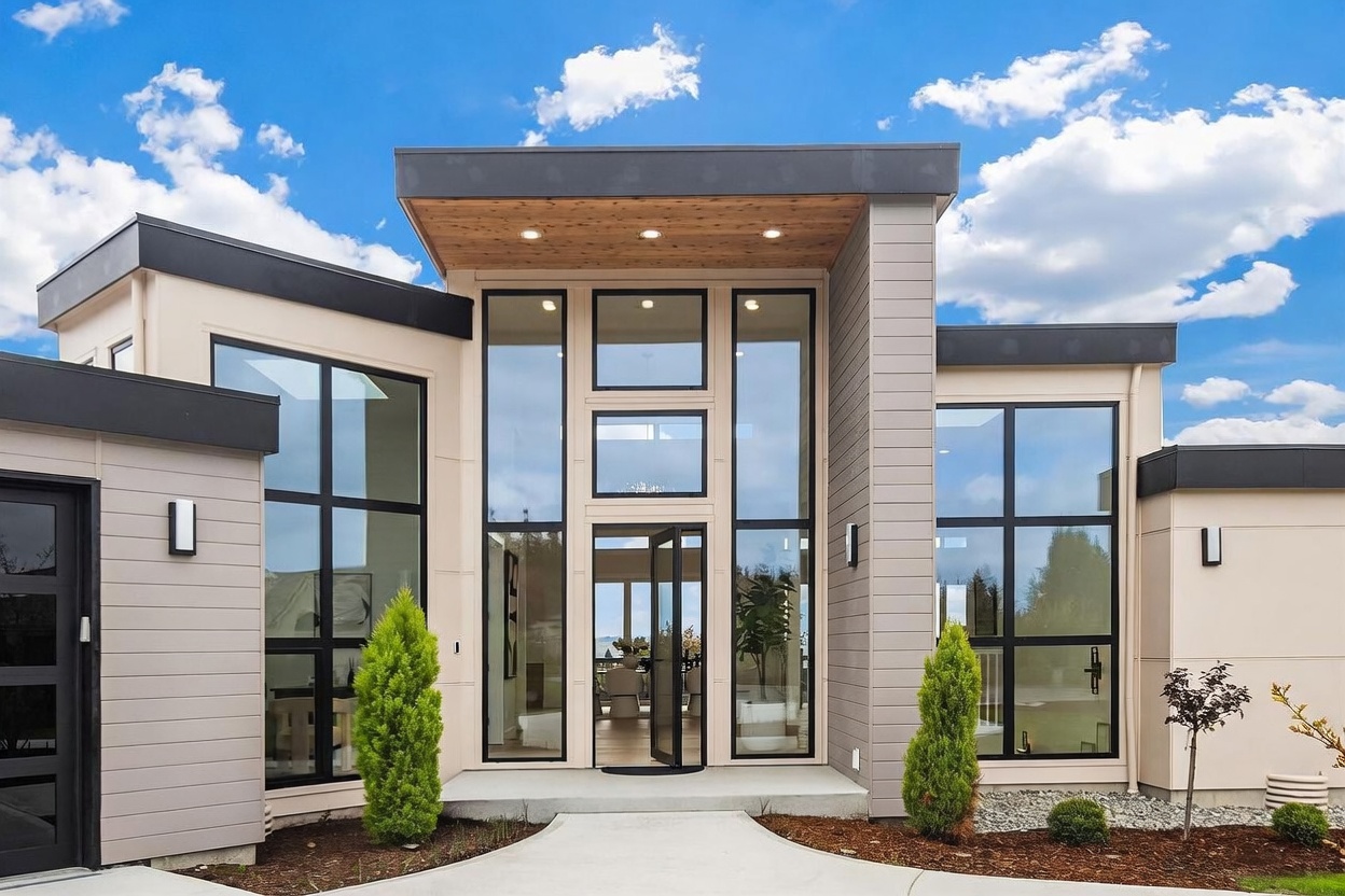 This modern home features a striking, symmetrical entryway with a prominent, flat-roofed portico accented by warm-toned wood soffits and recessed lighting. The facade is defined by clean horizontal siding and expansive, floor-to-ceiling windows that frame the central glass-paneled front door. Two manicured evergreen shrubs flank the concrete walkway, creating a polished and inviting curb appeal.