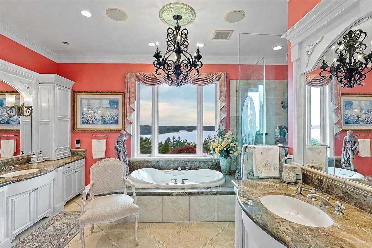 This luxurious primary bathroom features a central soaking tub positioned beneath a large window that offers expansive water views. The space is decorated with vibrant coral-colored walls, dual vanities with stone countertops, and an ornate chandelier hanging from the ceiling. A glass-enclosed shower and a decorative chair add to the elegant, traditional aesthetic of the room.