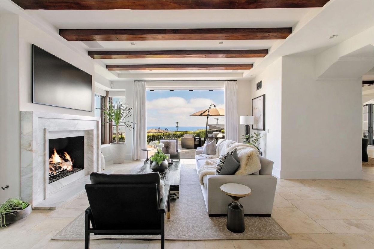 This elegant living room features exposed dark wood ceiling beams and a light-toned stone fireplace surround that serves as a focal point. A plush, light-colored sofa and a dark leather armchair are arranged on a textured area rug, creating a comfortable seating area. Large glass doors open fully to reveal a patio with an ocean view, seamlessly blending the indoor and outdoor living spaces.