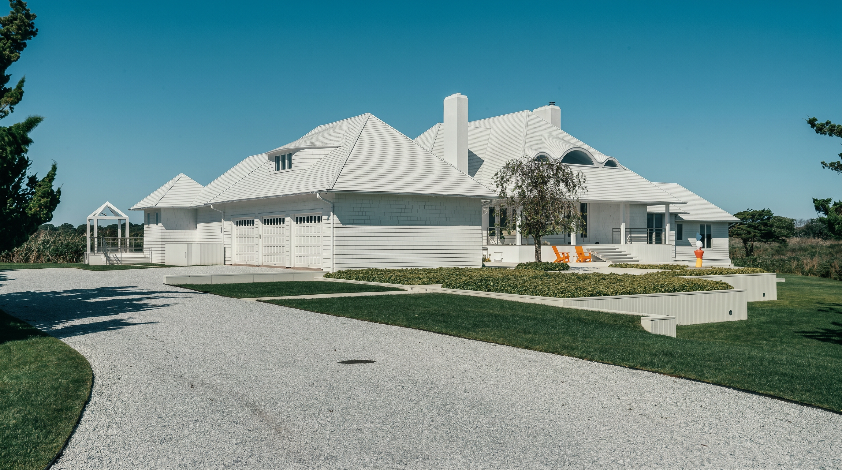 This expansive, white-shingled coastal residence features a multi-gabled roofline and a prominent chimney, set against a clear blue sky. A wide gravel driveway leads toward a three-car garage, while a manicured lawn and a raised garden bed with low-lying greenery frame the front entrance. The scene is captured from a low-angle perspective, emphasizing the home's architectural scale and its serene, open-air surroundings.
