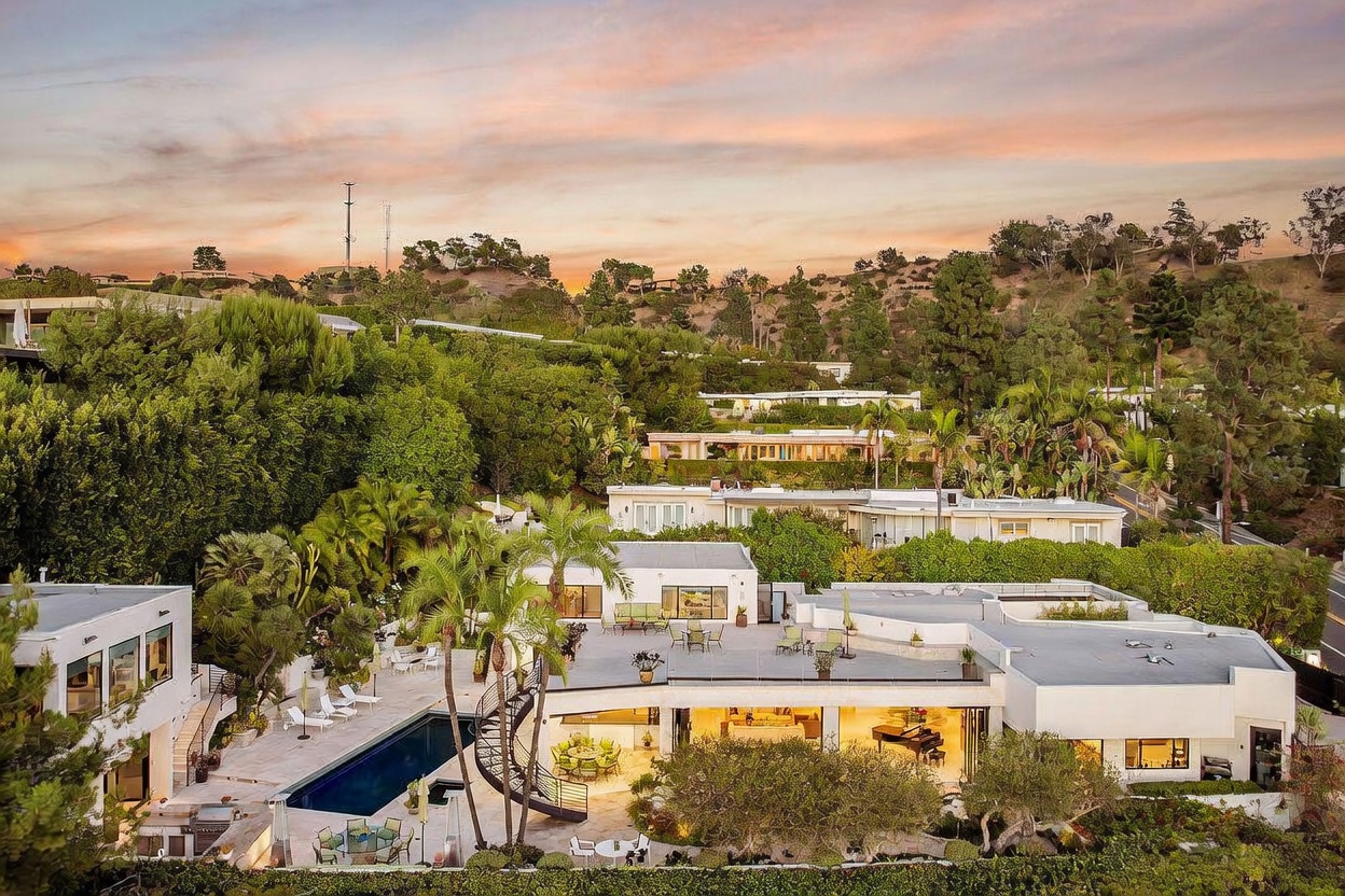 This aerial perspective captures a sprawling, modern residence nestled in a lush, hilly landscape during a vibrant sunset. The property features a multi-level layout with a large swimming pool, expansive paved patio areas, and a distinctive spiral staircase connecting the levels. The architecture emphasizes indoor-outdoor living with large glass walls that reveal a warm, inviting interior space.