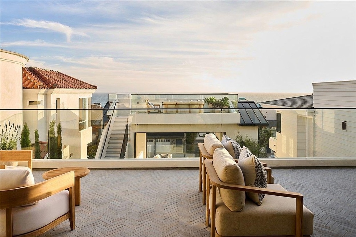 This elevated outdoor terrace offers a sophisticated lounge area with comfortable seating arranged on a herringbone-patterned brick floor. A sleek glass railing provides an unobstructed view of the ocean horizon and neighboring coastal homes. The space is designed for relaxation, capturing a serene atmosphere with a clear perspective of the surrounding architecture and the expansive sky.
