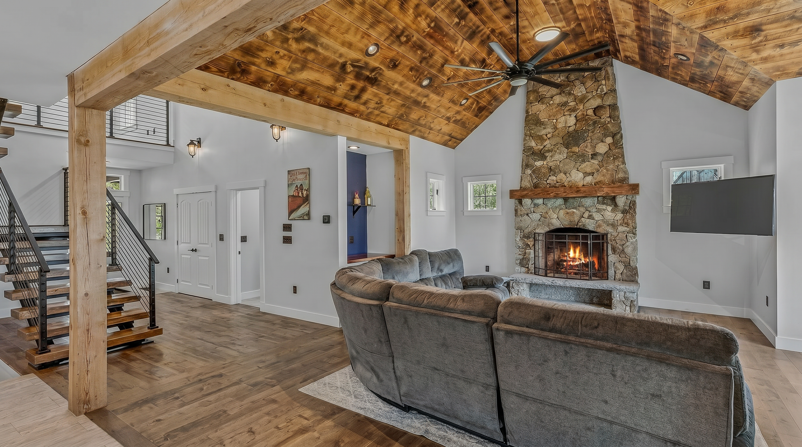This open-concept living area features a vaulted ceiling with exposed wood beams and a prominent stone-clad chimney breast housing a fireplace. A large, plush gray sectional sofa faces the fireplace and a wall-mounted television, while a staircase with metal railings and wood treads adds a modern industrial touch to the space. The room is finished with warm-toned wood flooring and neutral walls, creating a cozy yet spacious atmosphere.