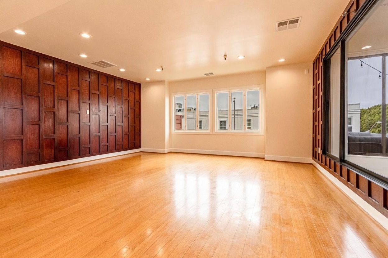 This spacious living room features light-toned wood flooring and a prominent accent wall covered in dark, grid-patterned wood paneling. Large windows on the back wall provide ample natural light, while a sliding glass door on the right opens to an outdoor space. The room has a clean, minimalist aesthetic with recessed lighting throughout, creating an open and airy atmosphere.