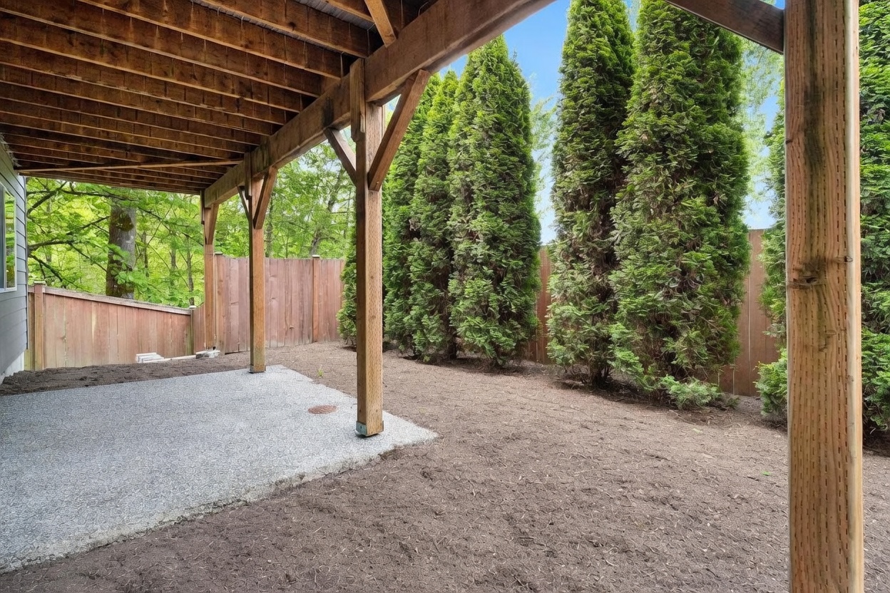 This image captures a covered patio area situated beneath a wooden deck structure, looking out toward a private backyard. The space features a textured concrete patio surface and is bordered by a wooden privacy fence and a row of tall, mature evergreen trees. The perspective is from under the deck, emphasizing the transition between the sheltered outdoor living space and the open, landscaped yard.