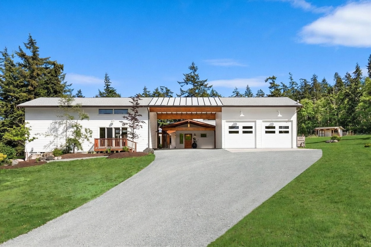 This modern single-story home features a crisp white horizontal siding exterior and a distinctive breezeway connecting the main living area to a two-car garage. A wide gravel driveway leads up to the central entryway, which is sheltered by a wood-accented roofline. The property is set against a backdrop of lush evergreen trees under a clear blue sky, creating a serene and inviting curb appeal.