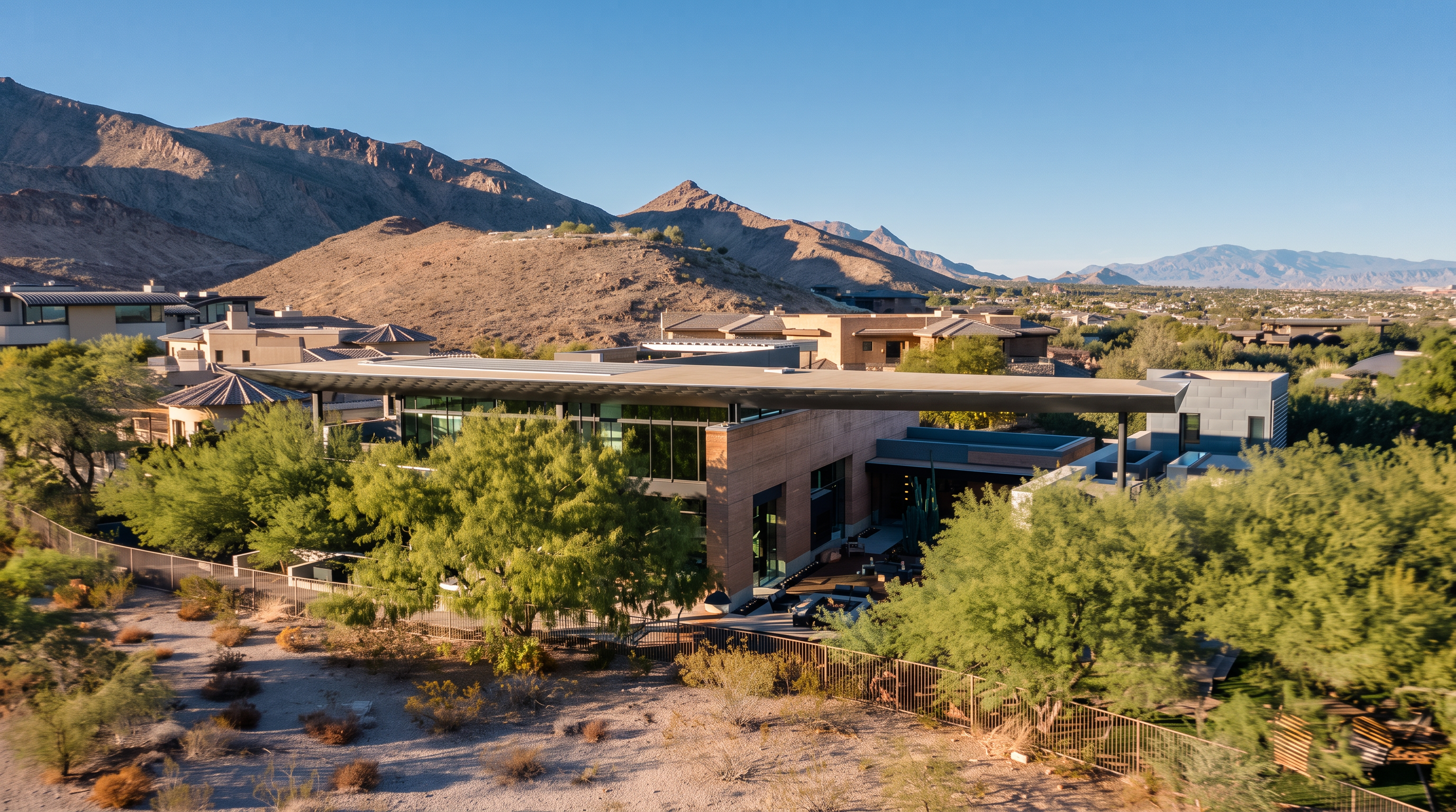 This elevated perspective captures a contemporary luxury residence set against a dramatic desert mountain backdrop. The home features a prominent, expansive flat roofline that extends over a glass-walled living space, blending modern architectural design with the rugged natural landscape. A fenced yard with native desert vegetation surrounds the property, emphasizing a seamless connection between the sophisticated structure and its arid environment.