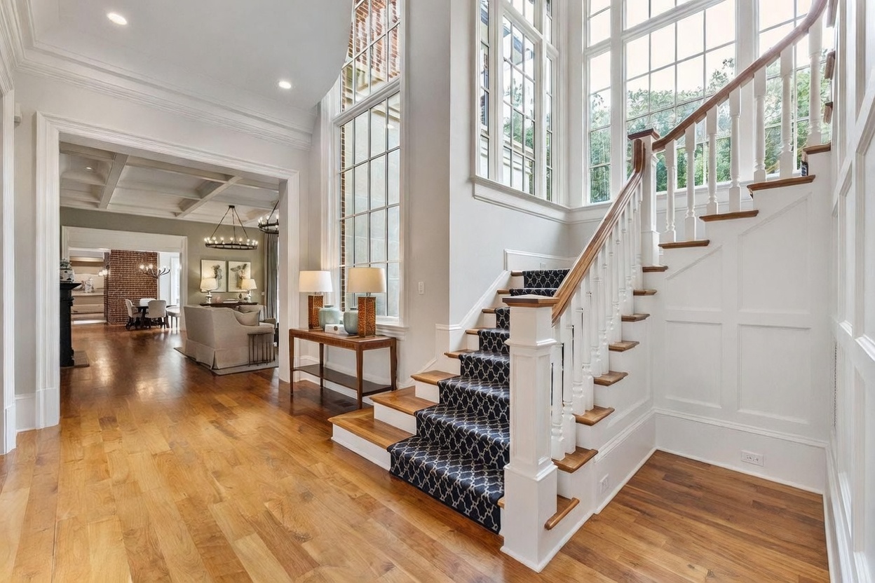 This grand foyer features a prominent staircase with a patterned runner and a white wooden railing, leading to an upper level. To the left, an open transition leads into a living area with light-toned furniture and a view of a dining space with a brick accent wall. Large, tall windows flood the space with natural light, highlighting the warm-toned wood flooring and the elegant, airy architectural design.