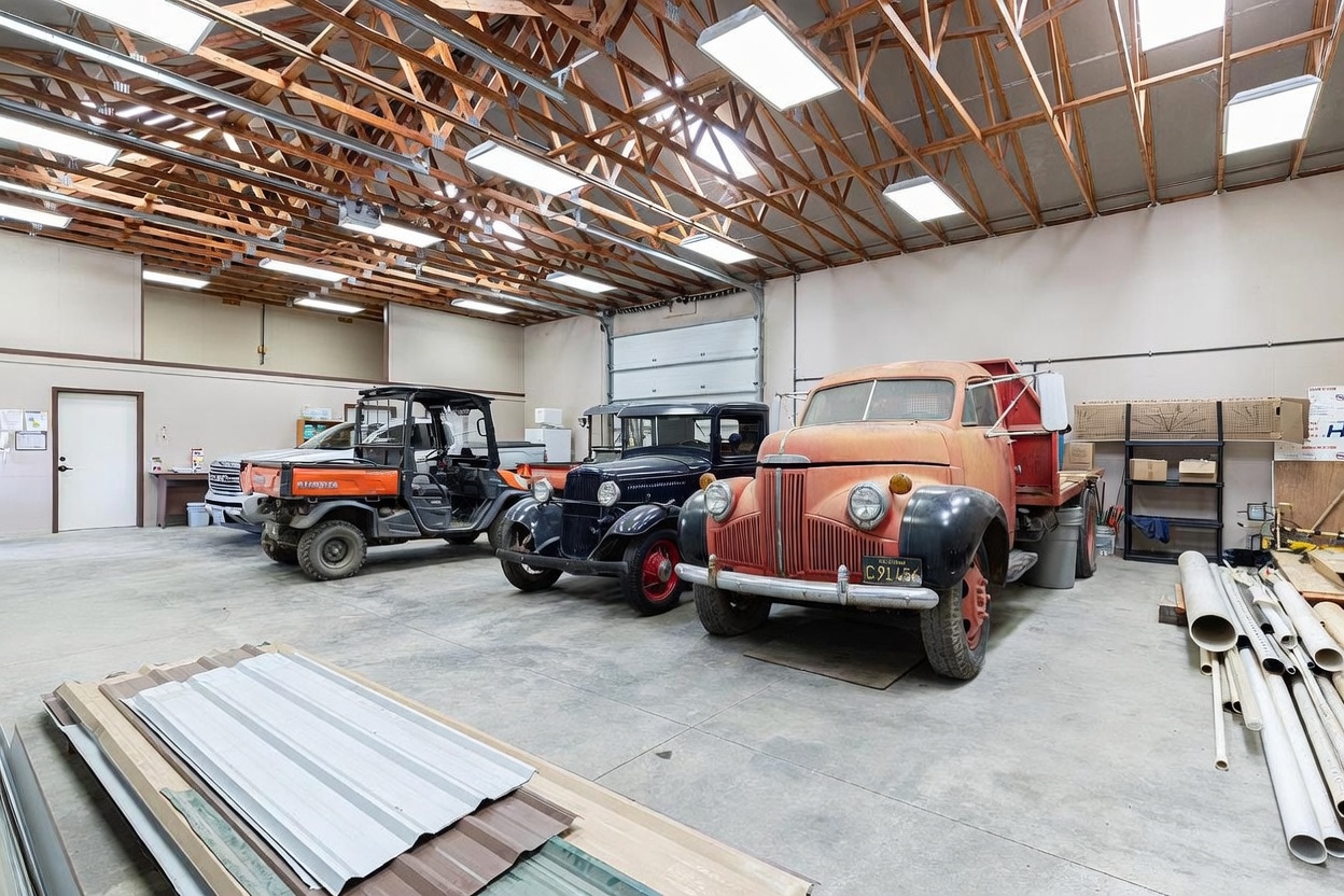 This spacious, high-ceilinged garage features exposed wooden trusses and multiple overhead lights, providing ample room for storage and vehicle maintenance. The concrete floor houses a diverse collection of vehicles, including a vintage dump truck, a classic sedan, and a modern utility vehicle. The wide-open layout and industrial aesthetic make it a versatile space for hobbyists or professional use.