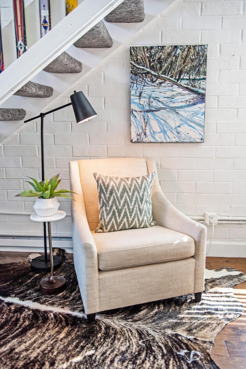 This cozy reading nook is situated beneath a staircase, featuring a neutral-toned armchair accented with a patterned throw pillow. A modern black floor lamp stands beside a small pedestal table holding a potted plant, all resting on a textured animal-hide rug. A piece of winter-themed landscape art hangs on the white painted brick wall, creating a warm and inviting atmosphere.