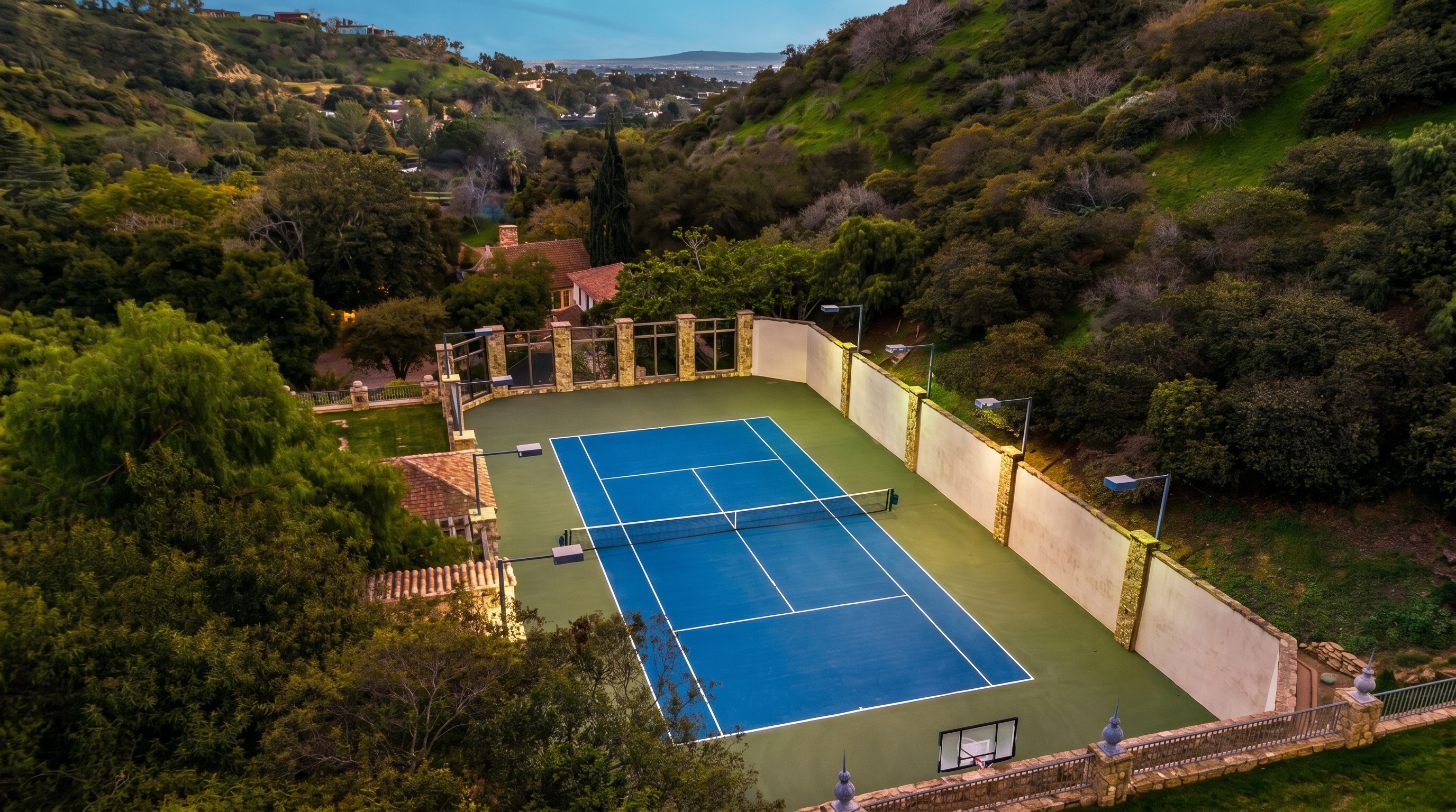 This high-angle aerial shot captures a private, professional-grade tennis court nestled into a lush, hilly landscape. The court features a vibrant blue playing surface with a basketball hoop at one end, surrounded by high retaining walls and mature trees that provide a sense of seclusion. The perspective emphasizes the property's integration into the natural terrain, highlighting a premium outdoor recreational amenity.
