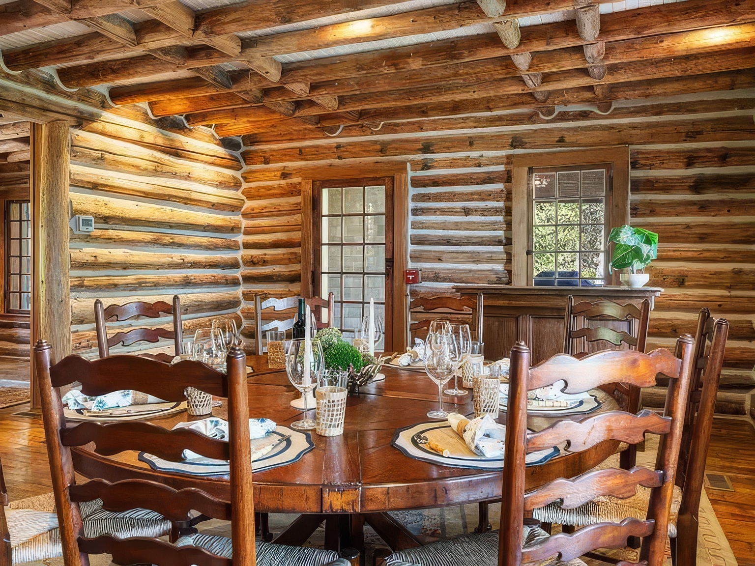 This rustic dining room features a large, round wooden table as the centerpiece, surrounded by traditional ladder-back chairs. The walls and ceiling are constructed from horizontal log timbers, creating a warm, cabin-like atmosphere. A glass-paned door and a window allow natural light to filter into the space, highlighting the rich wood textures and the formal table setting.