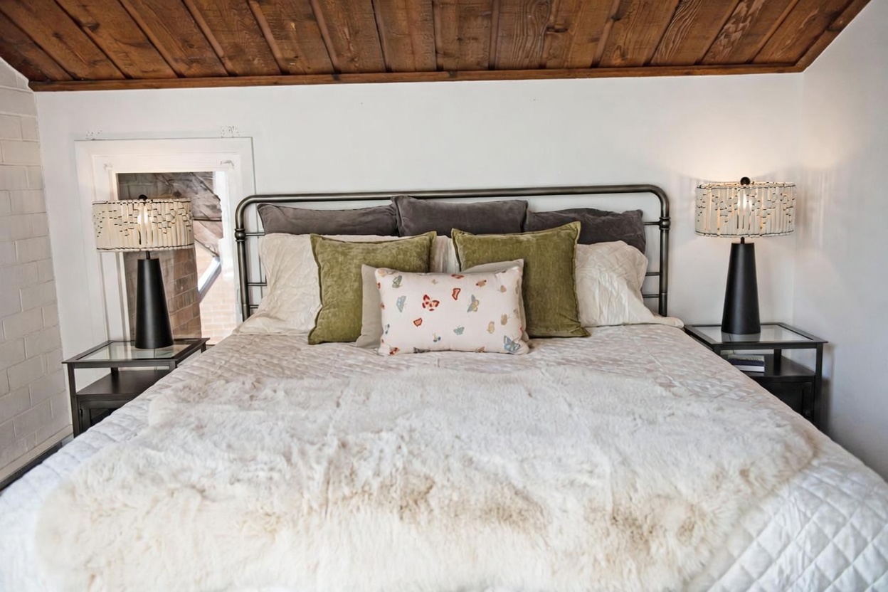 This cozy bedroom features a metal-framed bed topped with a plush, textured white throw blanket and a collection of decorative pillows in olive green and a butterfly-patterned accent. The room is framed by a rustic wood-paneled ceiling and white walls, with matching bedside tables and unique beaded lamps on either side. The perspective is a centered, eye-level shot that highlights the symmetrical arrangement and inviting atmosphere of the sleeping space.