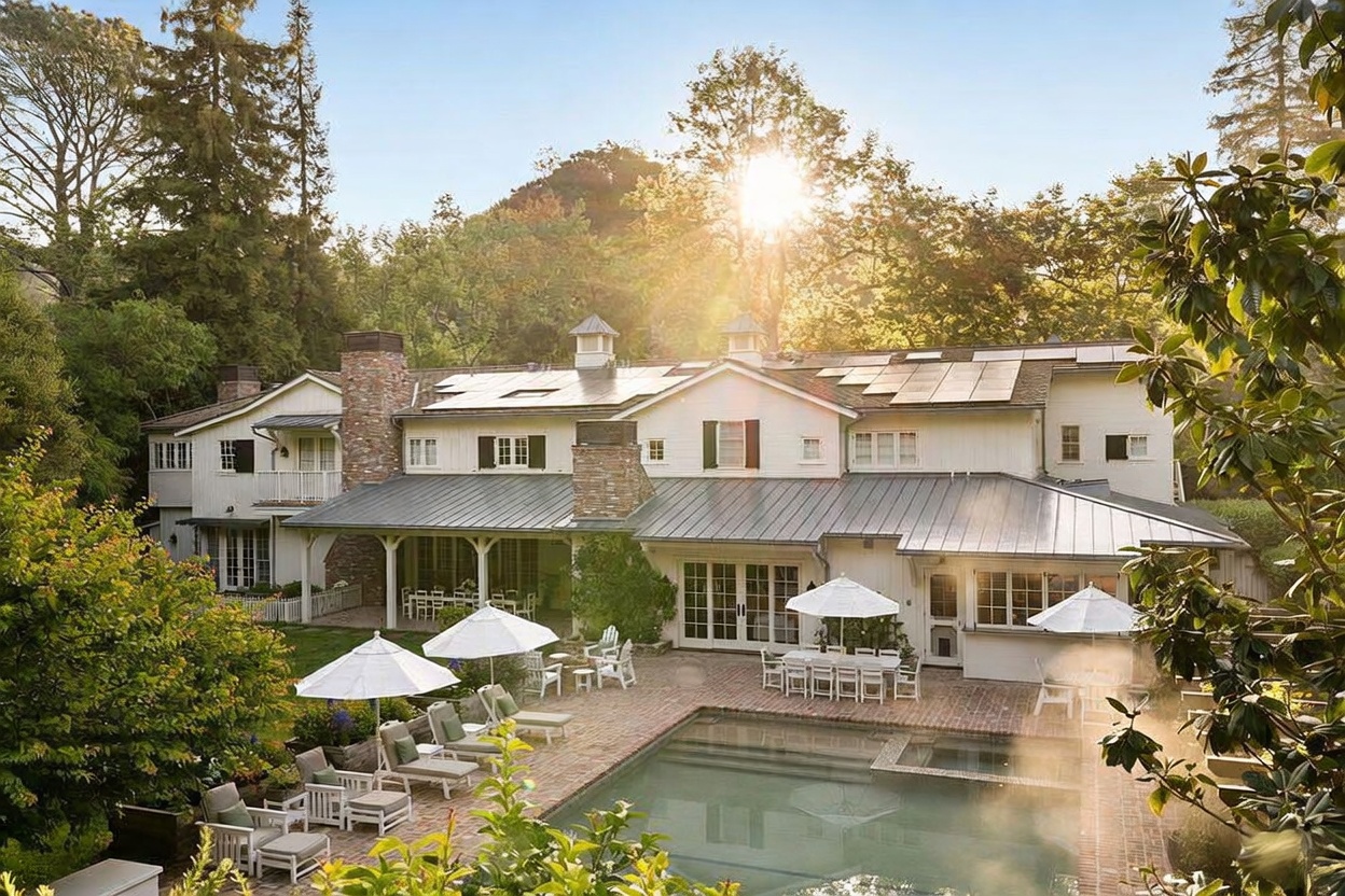 This expansive rear view of a traditional-style estate showcases a multi-gabled white home with a metal roof and solar panels. The property features a large brick patio surrounding a swimming pool, complete with multiple white umbrellas and outdoor seating areas. The scene is bathed in warm, golden-hour sunlight filtering through the surrounding mature trees, creating an inviting and serene atmosphere.