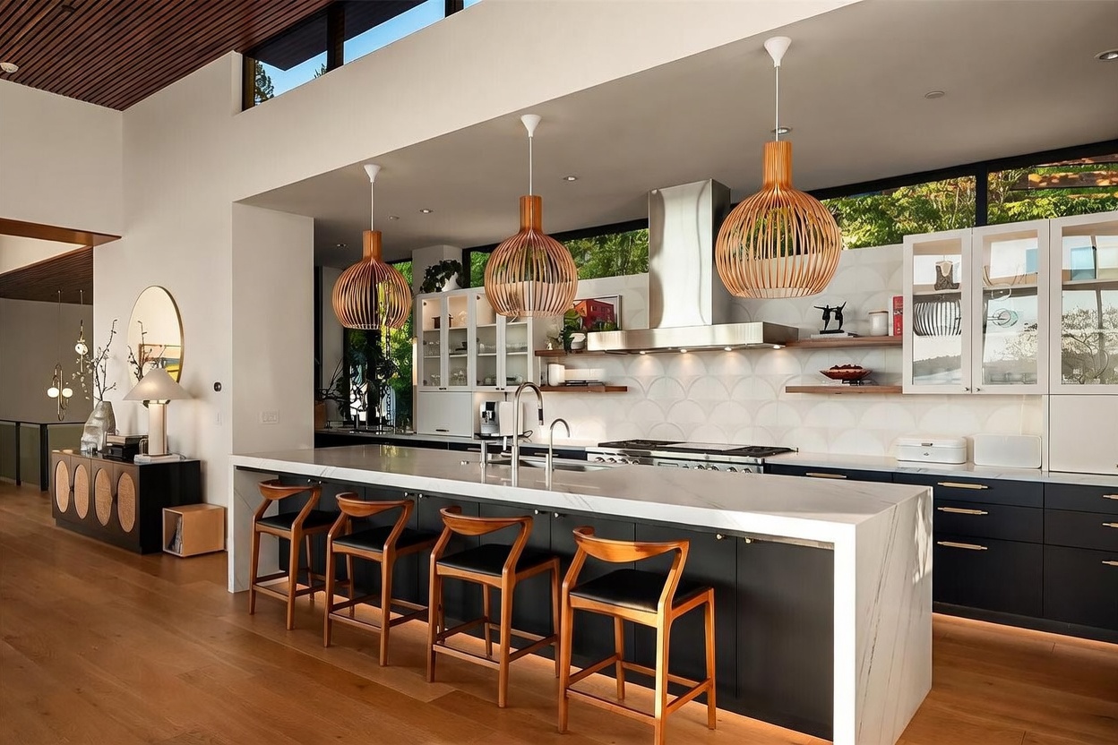 This modern kitchen features a large central island with a stone countertop and seating for four, complemented by dark cabinetry and light wood flooring. Three distinctive slatted pendant lights hang above the island, while a patterned tile backsplash and open shelving add visual interest to the cooking area. The space is brightened by clerestory windows that allow natural light to filter in, creating an open and airy atmosphere.
