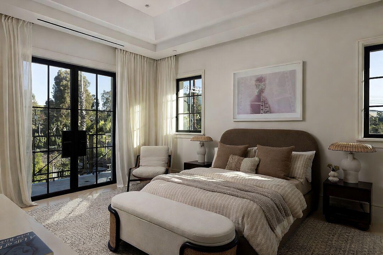 This serene primary bedroom features a neutral color palette with soft textures, including a plush upholstered bed and a matching bench at the foot. Large, black-framed glass doors open to a balcony, allowing natural light to fill the room, while a piece of framed art hangs above the headboard. The space is accented by two bedside tables with textured lamps, creating a sophisticated and tranquil atmosphere.