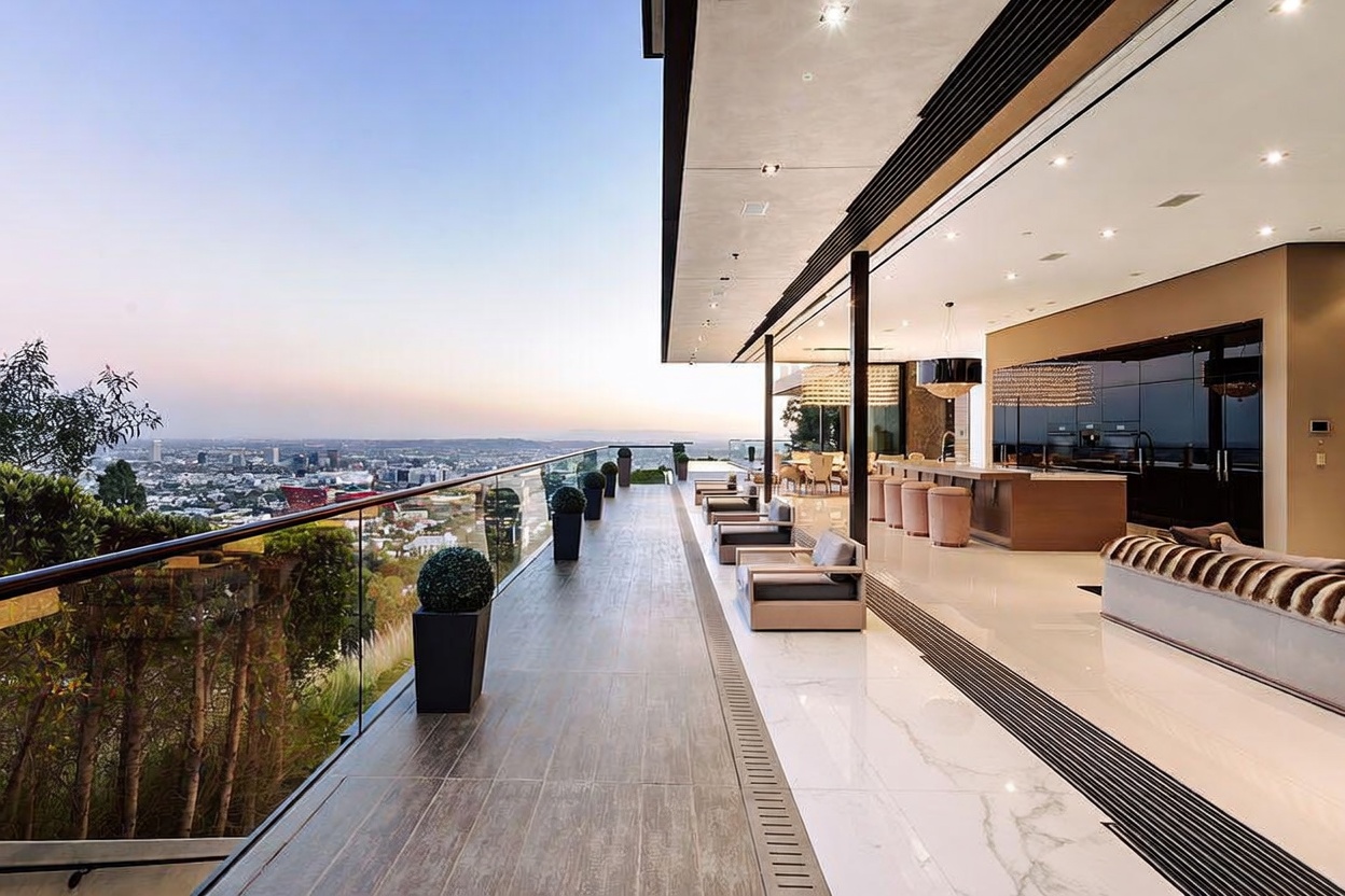 This expansive, modern balcony offers a seamless indoor-outdoor living experience with panoramic views of the city skyline at dusk. The space features sleek, contemporary outdoor furniture, a long kitchen island with bar seating, and a polished tile floor that transitions into the interior living area. The minimalist design and glass railing emphasize the dramatic height and luxury of this high-end property.