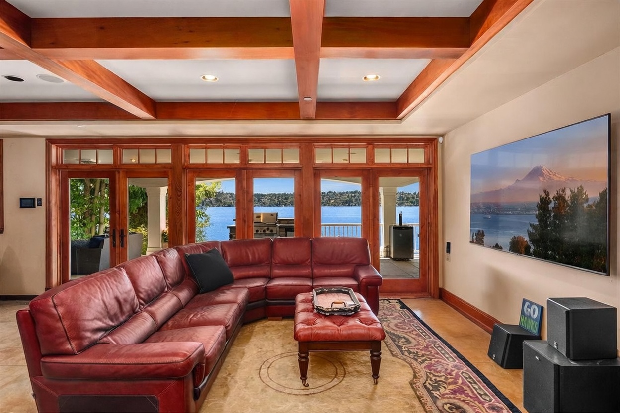 This spacious living room features a large, plush red leather sectional sofa and a matching ottoman centered on an ornate area rug. The room is defined by its striking coffered wood ceiling and a wall of floor-to-ceiling windows that provide a panoramic view of the lake and outdoor deck. A massive wall-mounted television dominates the side wall, creating an ideal space for entertainment in a luxurious, waterfront setting.