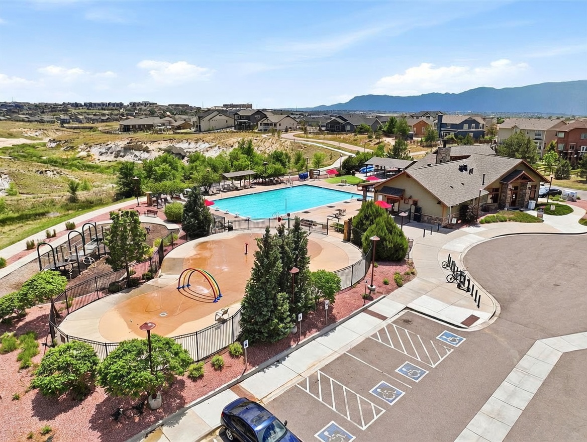 This high-angle aerial view captures a community recreation area featuring a large swimming pool, a splash pad with a rainbow-shaped water feature, and a clubhouse building. The facility is surrounded by landscaped grounds, paved walkways, and a parking lot with designated accessible spaces. The scene is set against a backdrop of suburban homes and distant mountains under a clear blue sky.