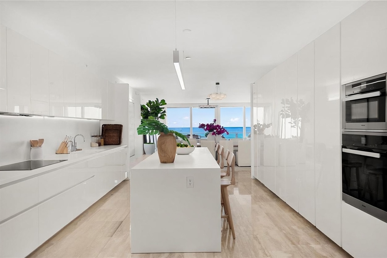 This sleek, modern kitchen features high-gloss white cabinetry and a central island with a light wood base, creating a bright and minimalist aesthetic. The space is anchored by a long, linear pendant light and opens directly toward a dining area with expansive, floor-to-ceiling windows that frame a stunning ocean view. The neutral color palette and clean lines emphasize a sophisticated, airy atmosphere perfect for contemporary coastal living.