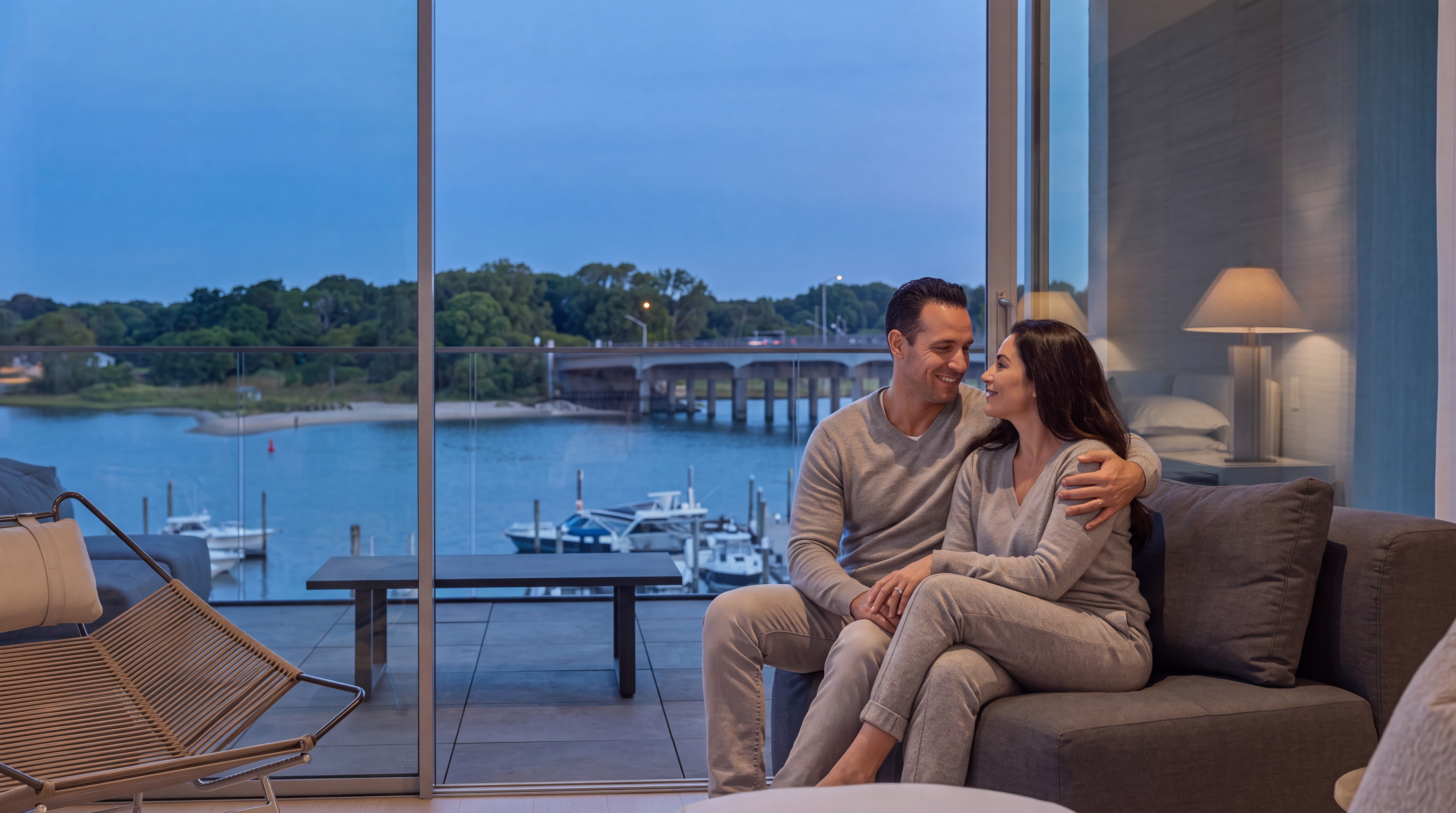 This living room features expansive floor-to-ceiling glass sliding doors that open onto a balcony with stunning waterfront and bridge views. The interior boasts a contemporary, minimalist aesthetic with clean lines, neutral tones, and comfortable chic furnishings. It offers an inviting and sophisticated ambiance, perfectly capturing the seamless transition between indoor luxury and picturesque outdoor scenery.