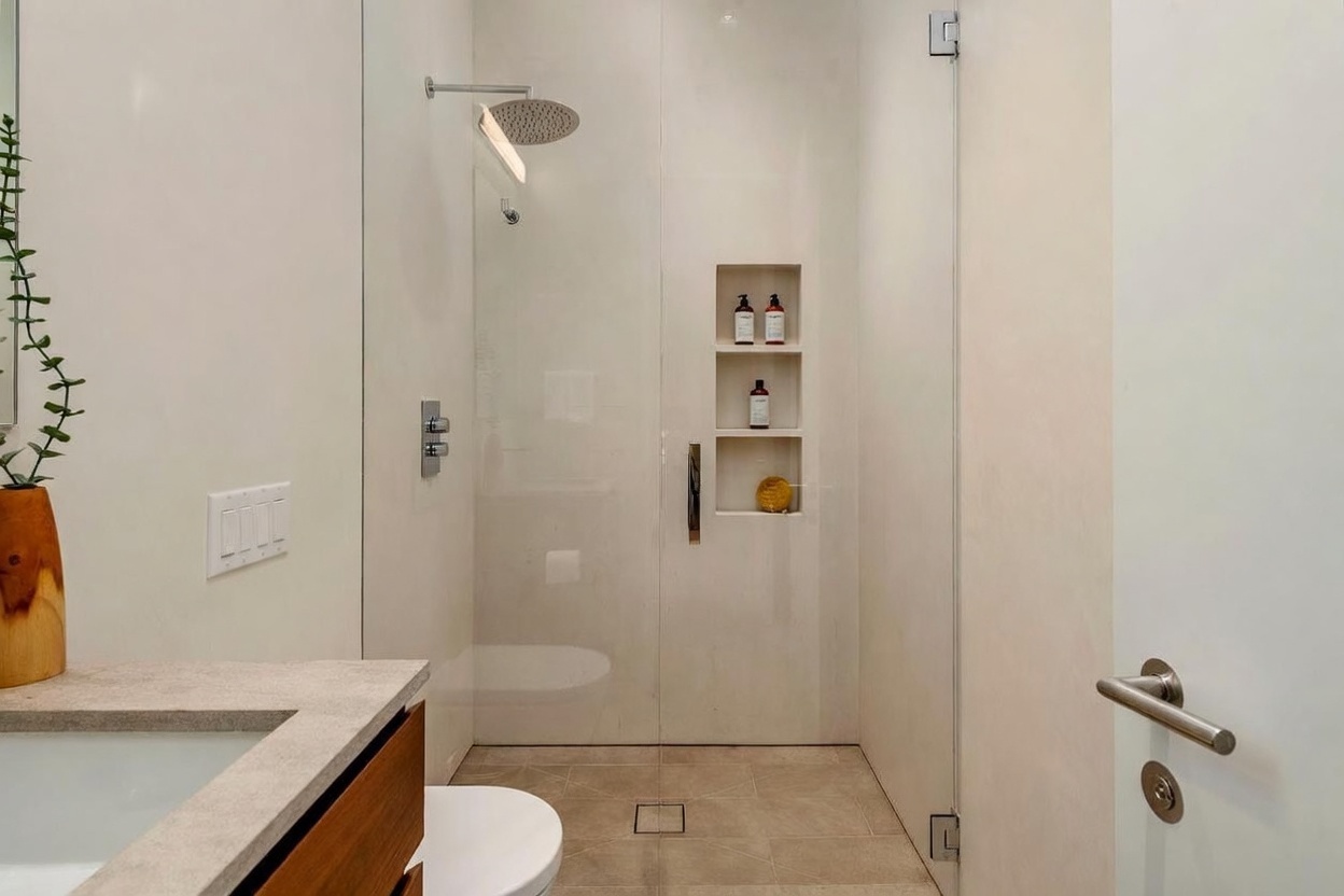 This modern bathroom features a walk-in shower enclosed by a glass panel, showcasing a rainfall showerhead and built-in recessed shelving for toiletries. To the left, a vanity with a light-toned stone countertop and a wood-accented cabinet provides a clean, minimalist aesthetic. The space is finished with neutral-toned floor tiles and a sleek, contemporary door handle, creating a bright and cohesive atmosphere.