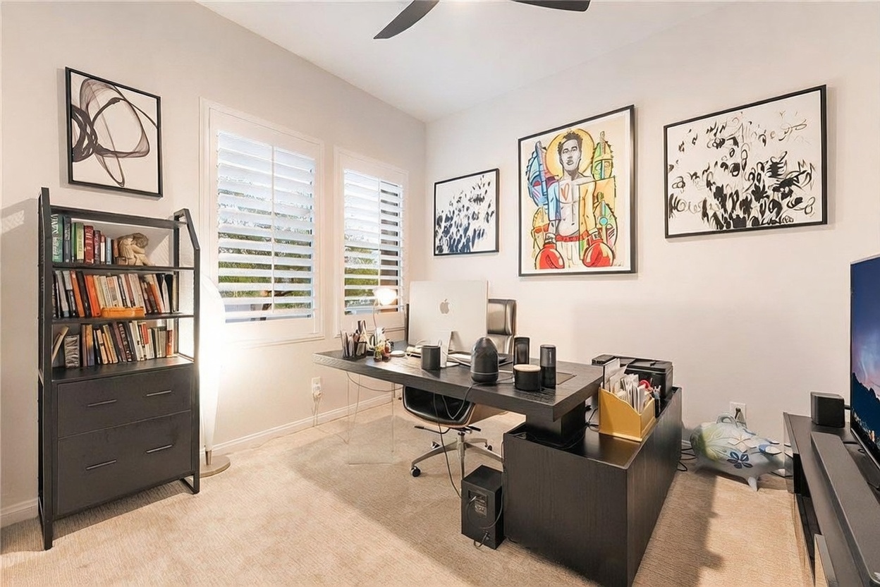 This home office features a dark wood desk and a matching bookshelf, creating a sophisticated and focused workspace. The room is decorated with vibrant, modern artwork on the walls and is illuminated by natural light filtering through white plantation shutters. A neutral-toned carpet and a ceiling fan complete the comfortable, professional atmosphere of this dedicated study area.