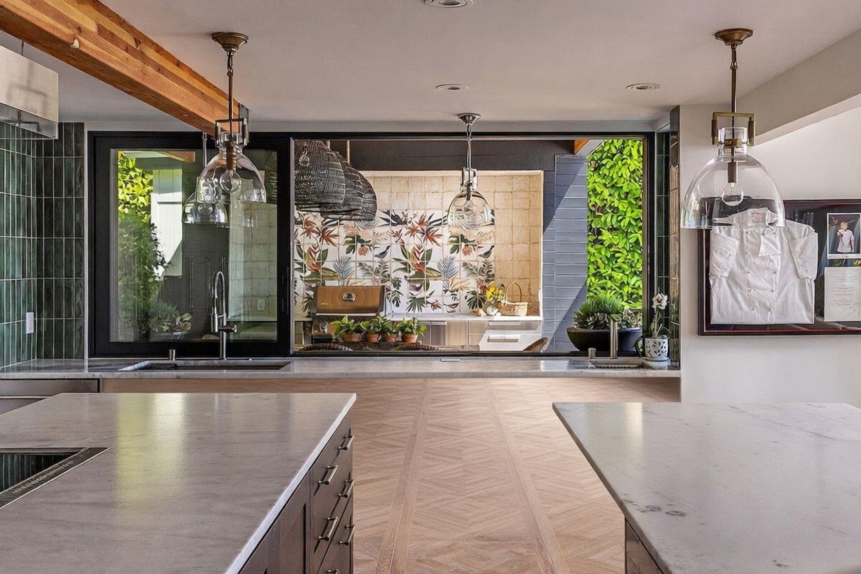 This modern kitchen features a large central island with light-toned stone countertops and dark cabinetry, complemented by a secondary island area. A large pass-through window connects the indoor kitchen to an outdoor cooking space, which is decorated with vibrant, patterned floral tiles. The space is illuminated by elegant pendant lighting and features a clean, sophisticated design with a seamless transition to the exterior.