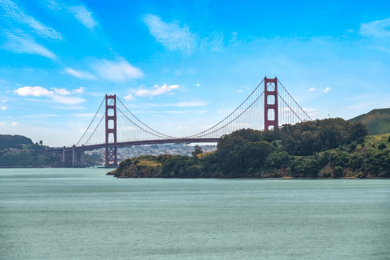 This wide-angle landscape shot captures the iconic Golden Gate Bridge spanning across the bay, framed by lush green hills in the foreground and a bright blue sky with scattered clouds. The perspective is taken from a distance, emphasizing the scale of the bridge against the natural coastal environment. The calm, turquoise water in the foreground provides a serene contrast to the structural complexity of the bridge towers and suspension cables.