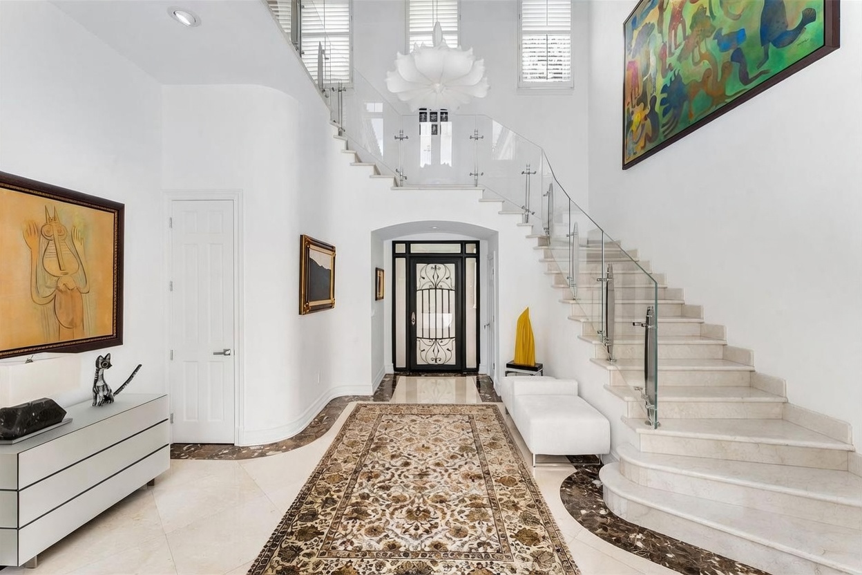 This grand entryway features a sweeping staircase with a glass railing and stone steps, leading to an upper landing. The space is characterized by high ceilings, a large decorative chandelier, and a long, ornate area rug centered on the polished stone floor. Art pieces adorn the walls, and a white console table sits to the left, creating a sophisticated and welcoming atmosphere.