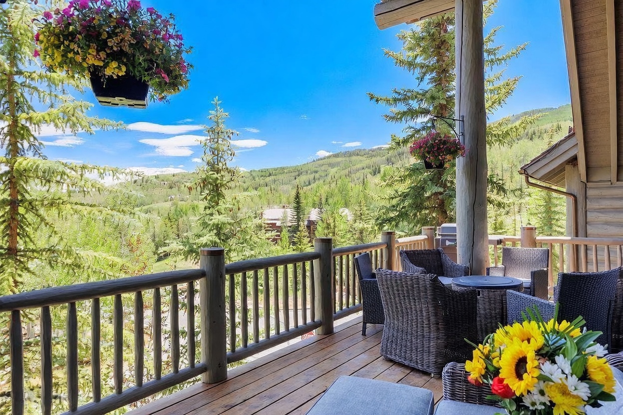 This elevated wooden deck offers a serene outdoor living space with panoramic views of a lush, forested mountain landscape. The area is furnished with comfortable wicker seating and a small table, accented by vibrant hanging flower baskets and a bouquet of sunflowers in the foreground. The rustic log-style railing and natural wood decking create a warm, inviting atmosphere perfect for enjoying the scenic surroundings.