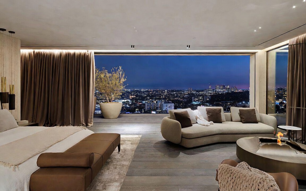 This luxurious primary bedroom features an open-concept design that seamlessly transitions to a spacious outdoor terrace, offering a breathtaking panoramic view of the city skyline at dusk. The room is elegantly furnished with a plush, curved sofa, a modern circular fire pit, and a sophisticated bed, all set against a backdrop of warm, ambient lighting and floor-to-ceiling glass walls. The aesthetic is ultra-modern and high-end, emphasizing a serene, resort-like atmosphere.
