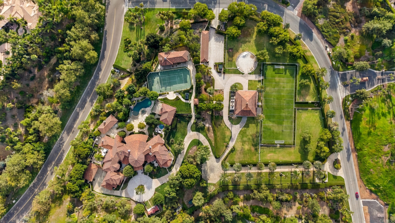 This aerial view showcases an expansive estate featuring a large Mediterranean-style home with a red tile roof, a swimming pool, a tennis court, and a soccer field. The property is surrounded by lush landscaping, mature trees, and winding driveways, creating a sense of privacy and luxury. The overall impression is one of grandeur and exclusivity.