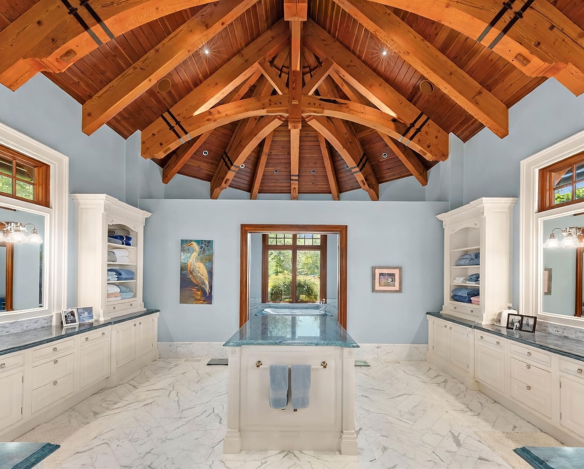 This luxurious primary bathroom features a dramatic vaulted ceiling with exposed timber beams and a central island topped with a blue-toned stone surface. The space is symmetrically designed with dual vanities, white cabinetry, and built-in shelving flanking a central doorway that leads to a soaking tub. The room is finished with light blue walls and patterned stone flooring, creating an elegant and serene atmosphere.