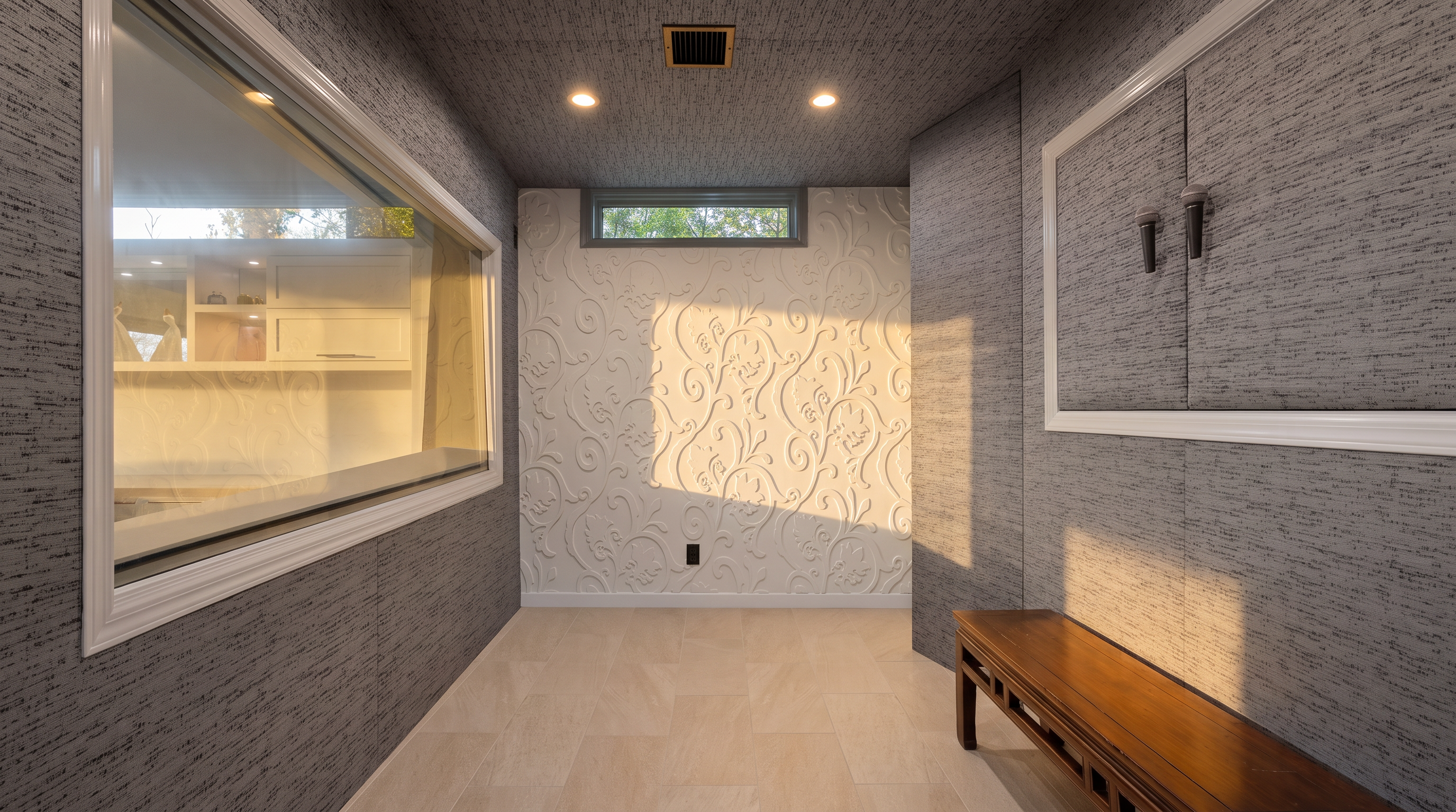 This unique interior space features a striking contrast between textured, dark gray wall coverings and a central wall adorned with a white, raised floral relief pattern. A long, rectangular window provides a view into an adjacent room, while a smaller, horizontal window sits above the decorative wall, allowing natural light to filter in. A dark wood bench sits on the light-toned tile floor, completing the sophisticated and modern aesthetic of this transitional area.