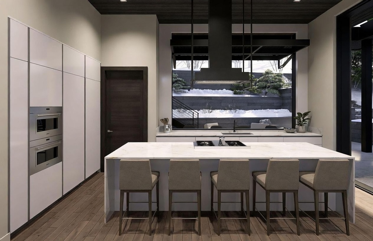 This modern kitchen features a large central island with a light-toned stone countertop and four bar stools, positioned in front of a sleek wall of white cabinetry and built-in stainless steel appliances. A dark-toned range hood hangs above the island, while a large window behind the sink provides a clear view of a snowy outdoor landscape. The space is defined by its clean lines, dark wood-toned ceiling, and warm wood flooring, creating a sophisticated and contemporary atmosphere.