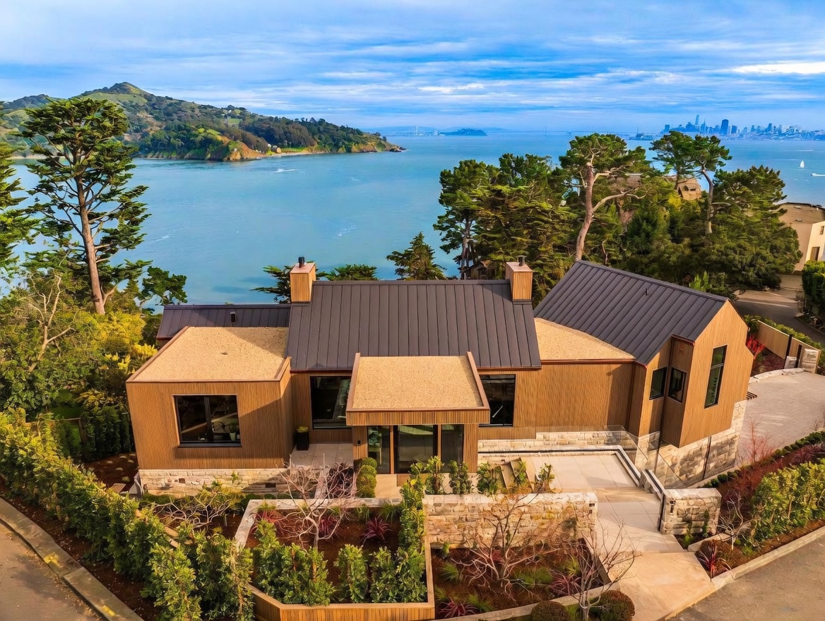 This elevated aerial perspective showcases a contemporary residence with a distinctive multi-gabled roofline and a blend of vertical wood siding and stone masonry. The home is situated on a scenic waterfront lot, offering expansive views of the bay and the distant city skyline. A tiered stone retaining wall and landscaped garden beds frame the front entrance, creating a sophisticated and inviting curb appeal.