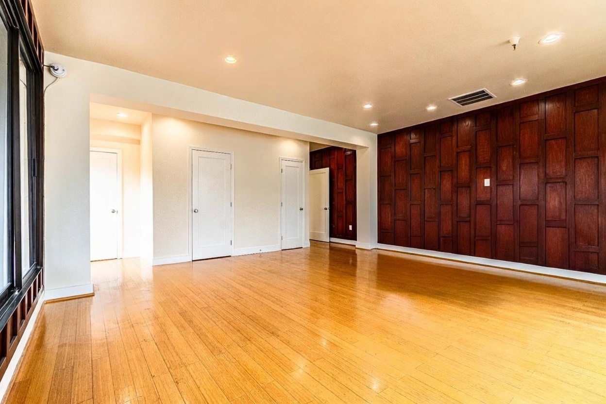 This spacious living area features warm-toned wood flooring and a prominent accent wall finished with dark wood paneling. The room is illuminated by recessed lighting and includes multiple white interior doors leading to adjacent spaces. The perspective captures the open layout, highlighting the contrast between the light-colored walls and the rich, textured wood feature wall.