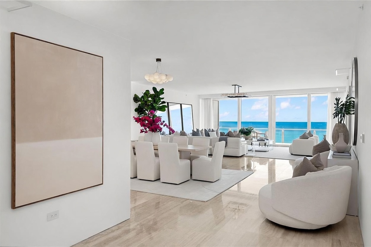 This bright and airy open-concept living and dining area features a sophisticated, neutral color palette with polished stone flooring. The space is anchored by a dining set and a plush seating area, all oriented toward expansive, floor-to-ceiling windows that frame a stunning ocean view. A large, minimalist abstract painting on the left wall adds a touch of artistic elegance to the clean, modern aesthetic.