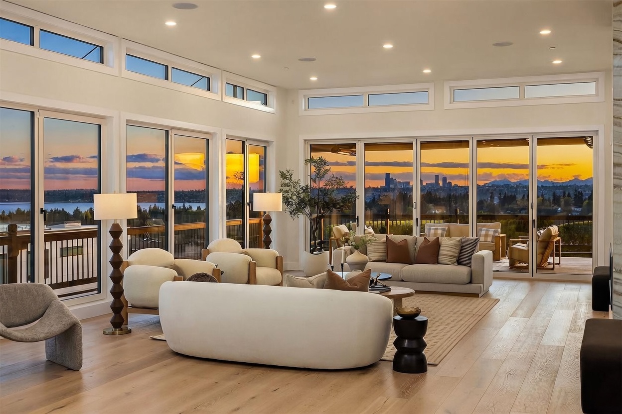 This expansive, light-filled living room features a contemporary open-concept design with light wood flooring and a neutral color palette. The space is defined by large, multi-panel glass doors that provide panoramic sunset views of the city skyline and water, creating a seamless transition to the outdoor deck. Furnished with a curved sofa, plush armchairs, and minimalist decor, the room offers a sophisticated and airy atmosphere perfect for entertaining.