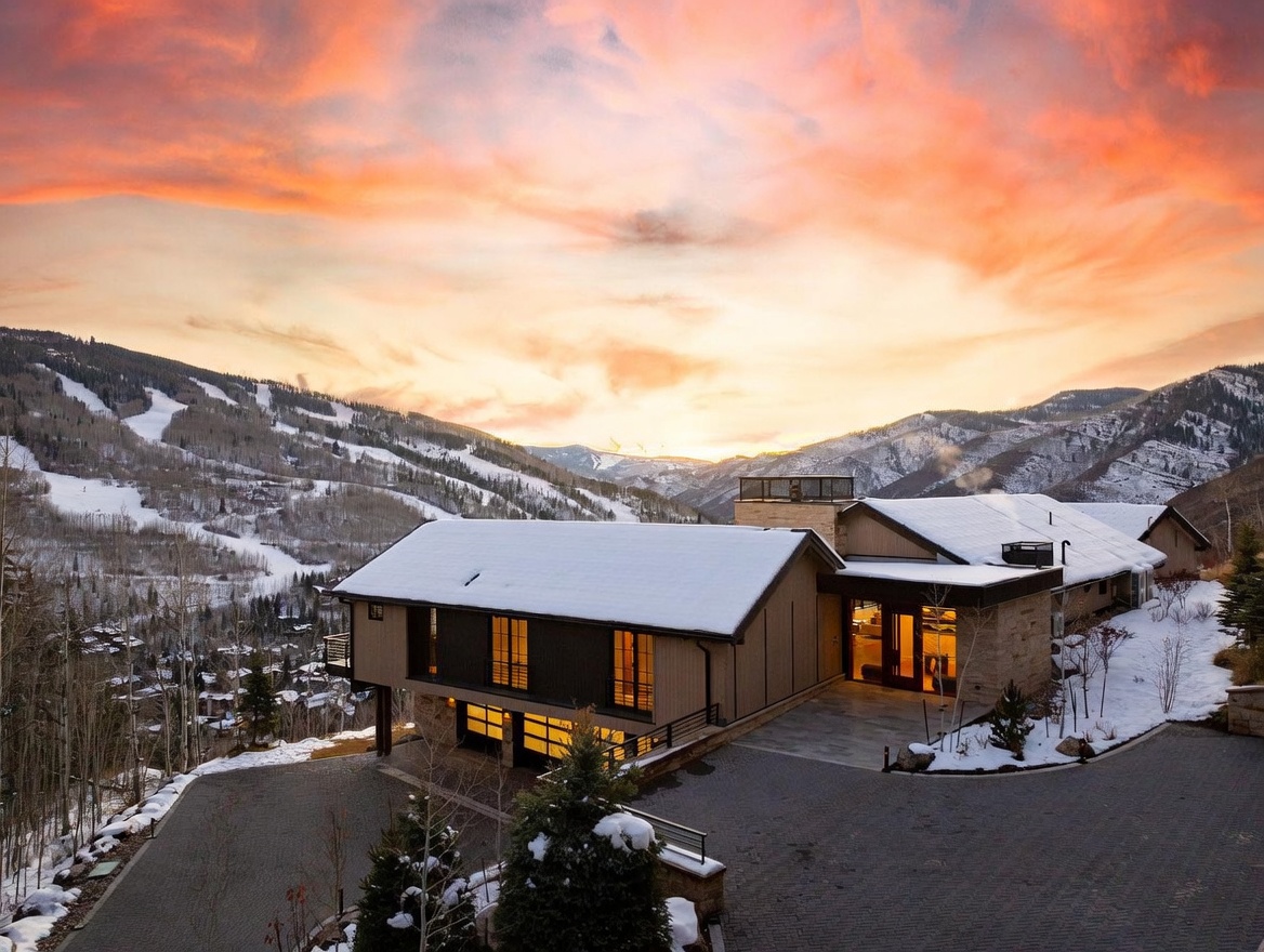 This elevated aerial perspective captures a modern mountain residence set against a dramatic sunset sky. The home features a multi-level design with a paved driveway, a multi-car garage, and expansive windows that overlook a snow-covered valley and ski slopes. The architecture blends contemporary lines with natural stone and wood textures, creating a sophisticated retreat in a high-altitude setting.