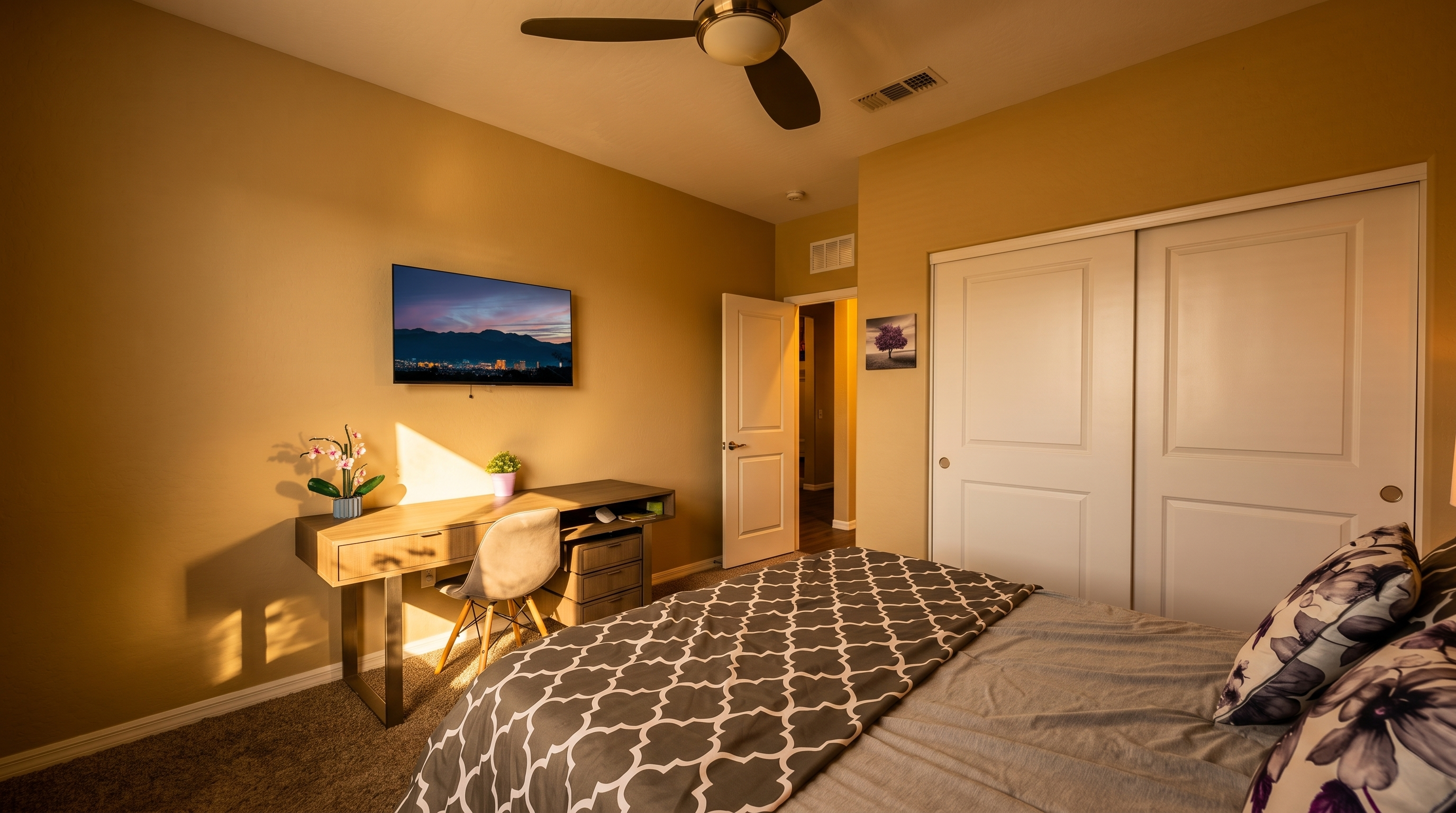 This bedroom features a neutral color palette with tan walls and gray carpeting, creating a calm and inviting atmosphere. The room is furnished with a bed dressed in a gray and white patterned comforter, a wooden desk with a gray chair, and a wall-mounted television. A ceiling fan provides air circulation, while white sliding closet doors and a doorway leading to a hallway complete the functional layout.