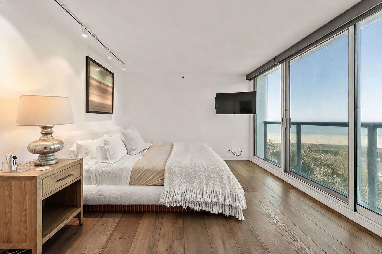This bright and airy primary bedroom features a minimalist aesthetic with light wood flooring and white walls. A plush bed with a textured white throw sits beside a wooden nightstand topped with a decorative lamp, while a wall-mounted television faces the bed. Large glass sliding doors provide an expansive view of the ocean and beach, filling the room with natural light.